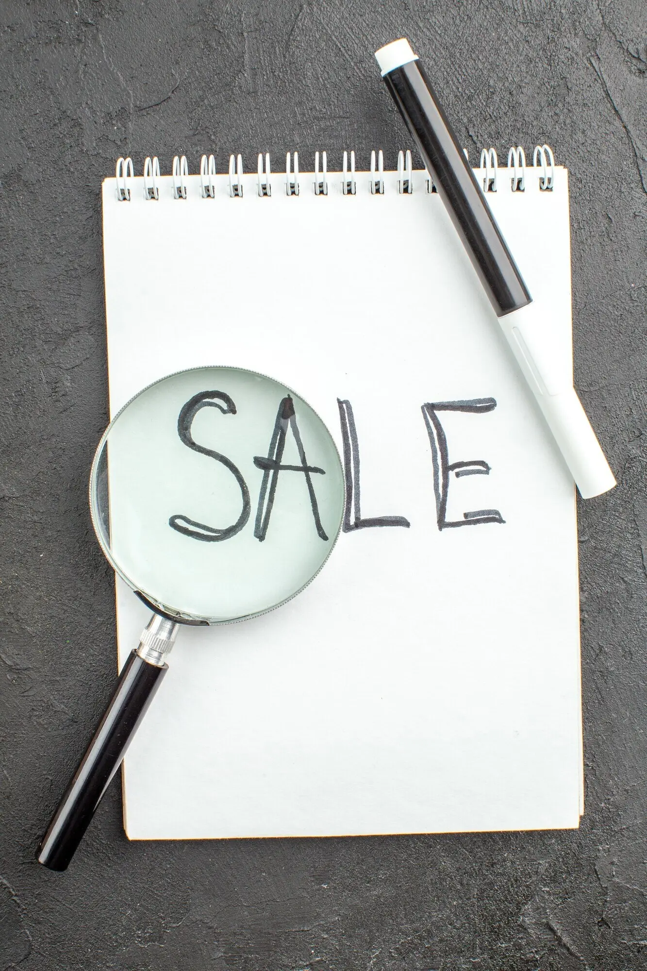 Ansicht von oben: „Sale“ auf einem Spiralnotizbuch geschrieben, Lupe und schwarzer Marker auf schwarzem Hintergrund.