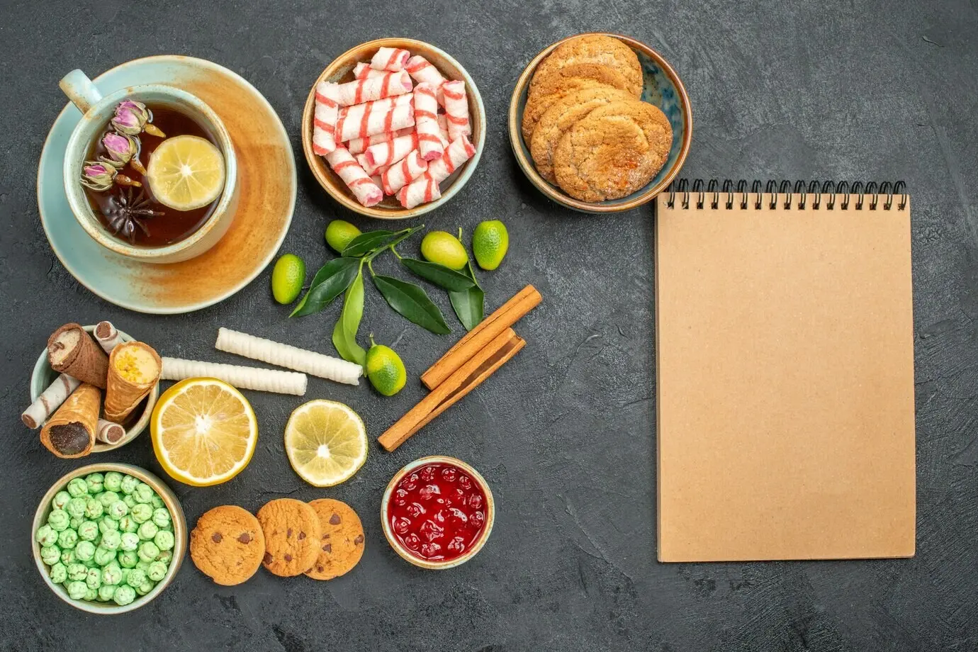 Nahaufnahme von oben mit einer Tasse Tee, einer Tasse Kräutertee, Bonbons, Waffeln, Marmelade, Sahne und einem Notizbuch.